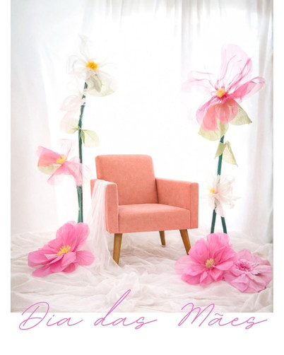 Cenário fotográfico para o Dia das Mães com uma poltrona rosa claro, flores gigantes de papel em tons de rosa e fundo branco. No canto inferior, a frase "Dia das Mães" em escrita cursiva rosa.