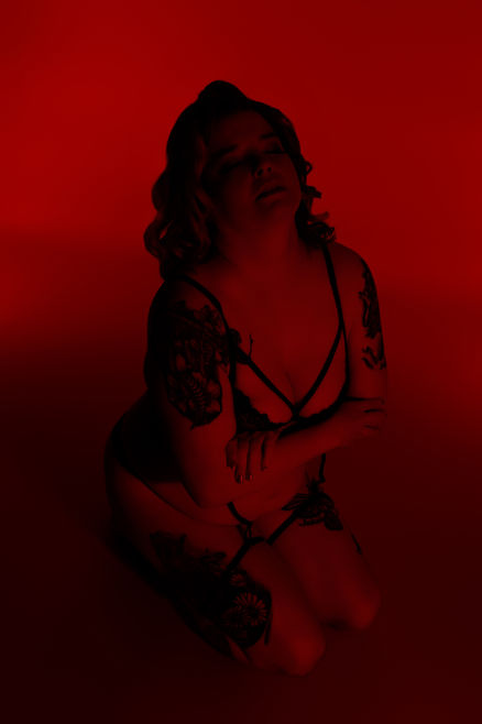 Moody red-lit boudoir portrait of a tattooed woman posing confidently in black lingerie. Luxury boudoir photography in York, PA.