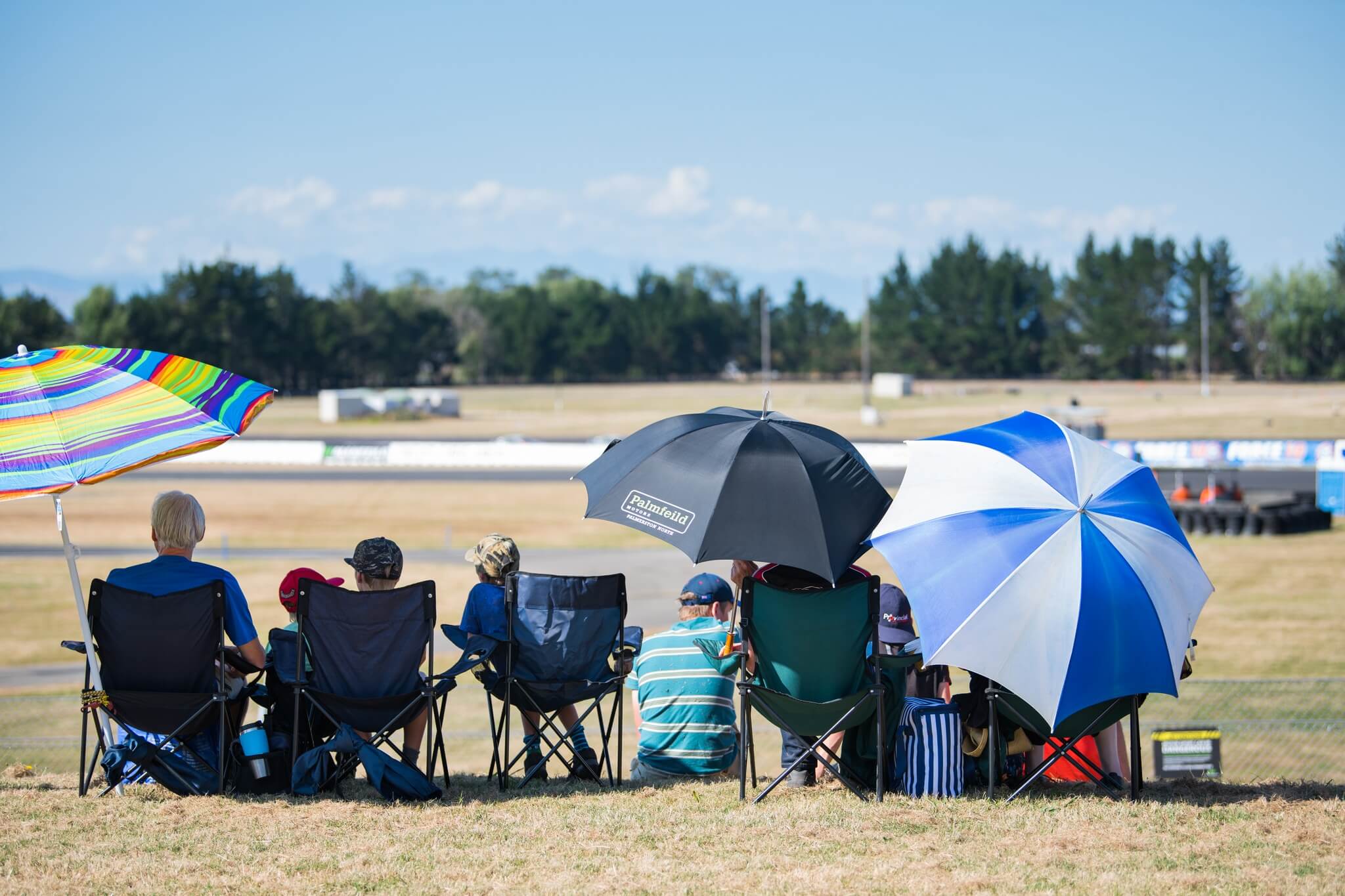 Spectators sitting in chairs under umbrellas watching an outdoor event on grass.