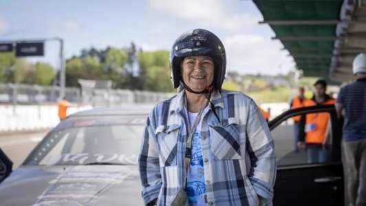 Smiling woman in helmet and plaid shirt beside race car on track.