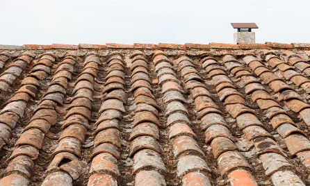 Weathered terracotta roof tiles with a small chimney in the background against a cloudy sky, creating a rustic and serene atmosphere.