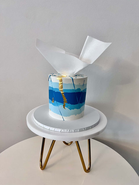 Single tier buttercream celebration birthday cake, white base with shades of blue colouring running concentrically around the cake. Decorated with edible gold leaf and moulded wafer paper topper