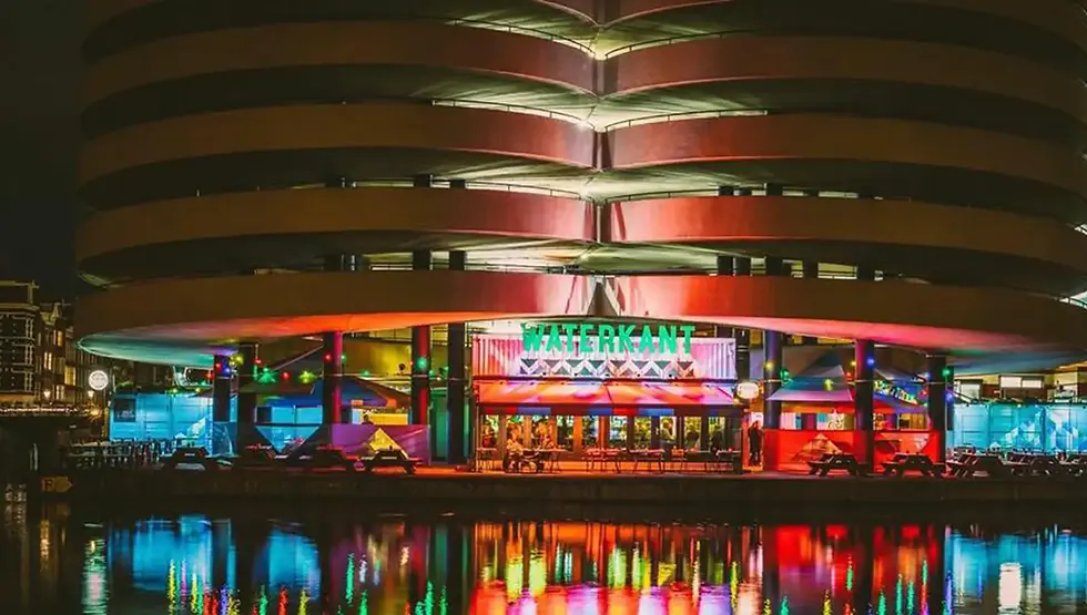 Colorful building at night with "WATERKANT" sign, reflecting in water. Vibrant lights illuminate the modern architecture, creating a lively mood.