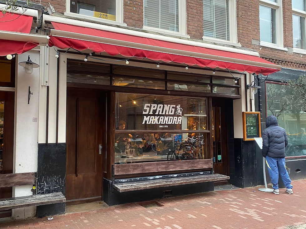 Street view of a café with red awnings. A person in a blue hoodie reads a menu outside. Sign reads "Spang Makandra." Warm interior.