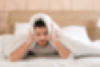 A man holds his blankets over his ears attempting to drown out noise so he can sleep.