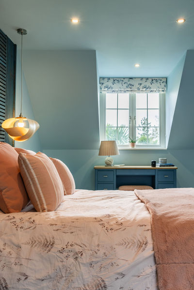 Soft blue bedroom with layered blush textiles and cosy styling. Bedroom window in backgroudn with patterned roman blind and soft light