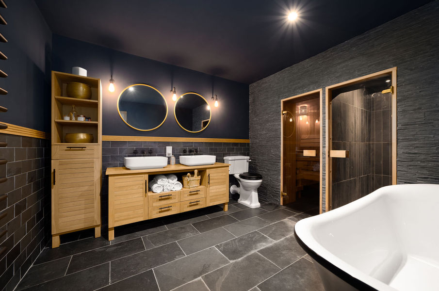 Luxury dark bathroom with twin oak vanity, round mirrors, freestanding bath and built-in sauna