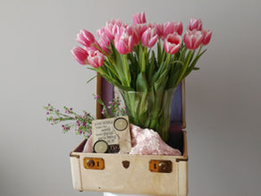 Pink tulips in a vase sit in an open suitcase with a plaque reading, "Some people make the world more special just by being in it."