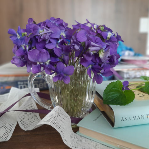 Bukiet fioletowych kwiatów w szklanym wazonie na tle książki "As a Man Thinketh". Obok białe i fioletowe wstążki. Przytulny nastrój.