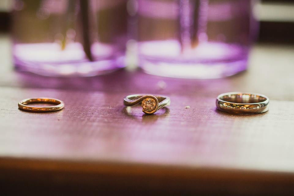 Three rings on a wooden surface, with a diamond ring centered between gold and silver bands. Blurred purple background adds a soft mood.