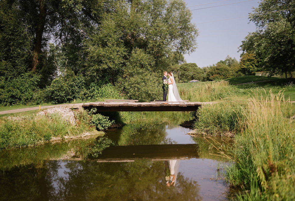 Bride and groom share an embrace on a wooden bridge over a stream, surrounded by lush greenery and clear sky, creating a romantic scene.