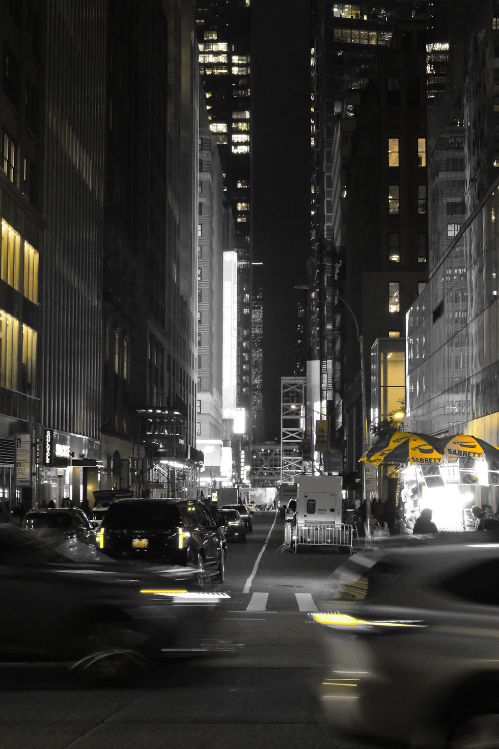 Black-and-white nighttime street scene in New York with yellow accents on umbrellas and car lights.
