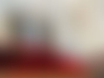 A bishop in ornate white and gold robes stands at a lectern in a church. Red votive candles and a blurred photo are in the foreground.