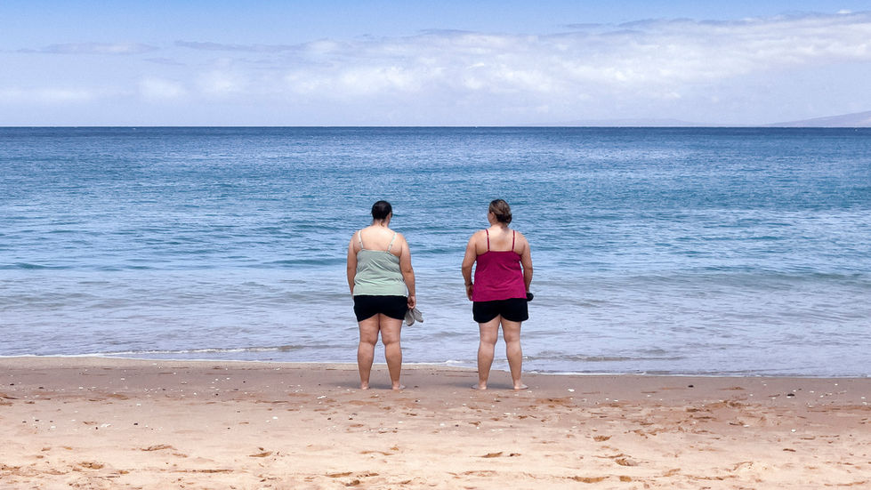 Two figures standing at the ocean's edge, seen from behind, facing the open Pacific.