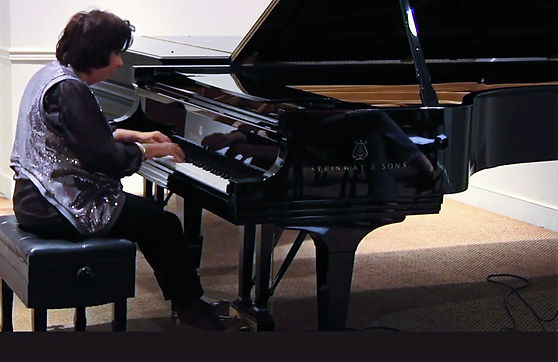Sedmara Zakarian Rutstein Performing on stage at Oberlin Conservatory Winter Concert. We see Sedmara playing a black grand piano. She is wearing a black blouse and trousers and a silver sequinned vest. We see Sedmara in profile as she is looking at the keys of the piano while playing.