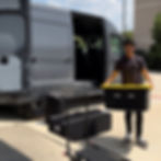 Man unloads a gray van with open door, surrounded by AV bags and a dolly on a sunny day.