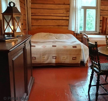 Kuvassa Punaisen Kaupan majoitustilaa. Etualalla vanha senkki, jonka päällä on lyhty. Takana näkyy parisänky ja vanha hirsiseinä. Sängyn vieressä on ikkuna, josta näkyy vehreitä puita.
