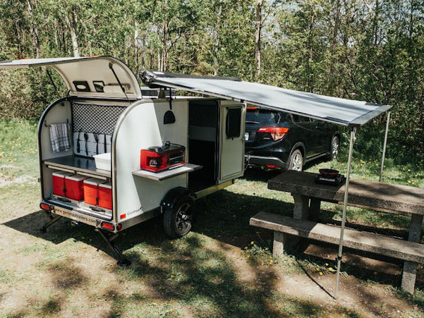 Roulotte Genimax installée en camping avec hayon arrière ouvert, cuisine extérieure, auvent déployé et espace repas aménagé.