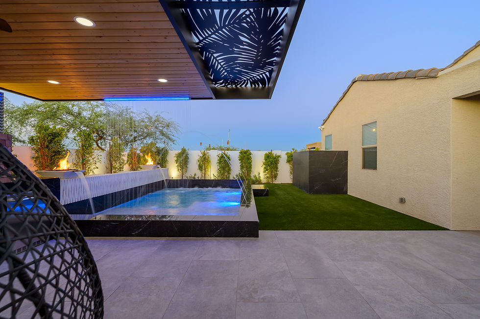 Modern backyard patio with glowing blue pool, waterfall, fire bowls, and wood pergola under dusk sky.
