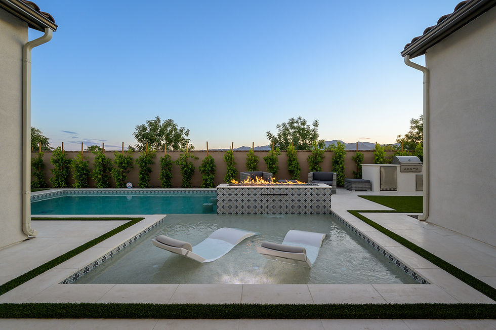 Modern backyard with a pool, fire feature, and two sun loungers. Greenery lines the fence. A BBQ grill is visible under a clear sky.