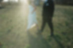 A couple walks on a grassy field with sunlight streaming. The woman wears a white dress, the man a black suit. Trees line the background.