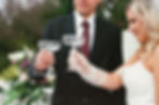 Bride and groom in formal attire clink champagne glasses, smiling. Outdoor setting with blurred greenery and flowers in the background.
