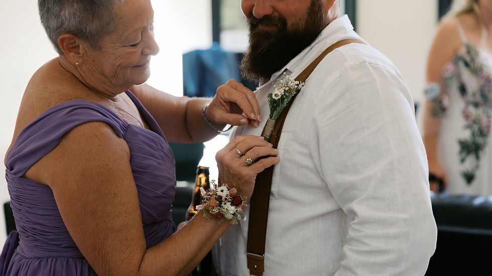 Woman in purple dress pins boutonniere on man's white shirt. Smiling in a softly lit room with floral decor. Warm, intimate mood.