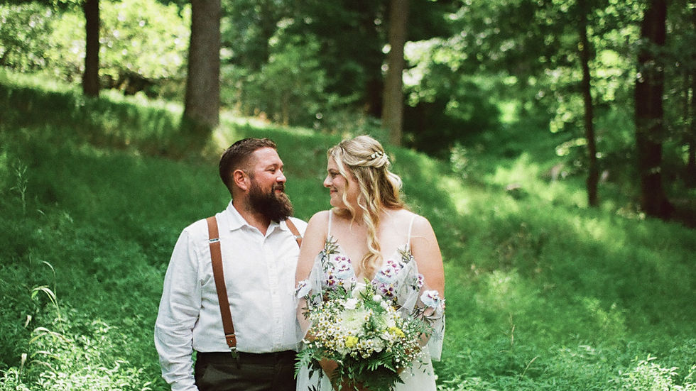 A couple stands in a lush green forest. The woman holds a bouquet, wearing a floral dress. Both smile at each other, creating a romantic mood. Shot on film