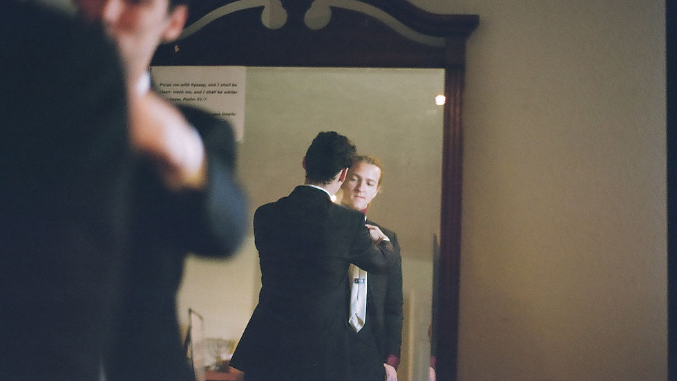 Two people in suits reflected in a mirror, one adjusting the other's tie. A small sign with text is visible. The setting is a dimly lit room.