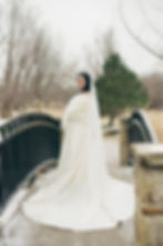 A woman in a white wedding dress stands on a bridge, wrapped in a fur stole. The background shows bare trees and a cloudy sky.