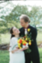 Bride and groom embrace, smiling at each other. Bride holds bouquet of yellow and red flowers. Lush green outdoor setting.