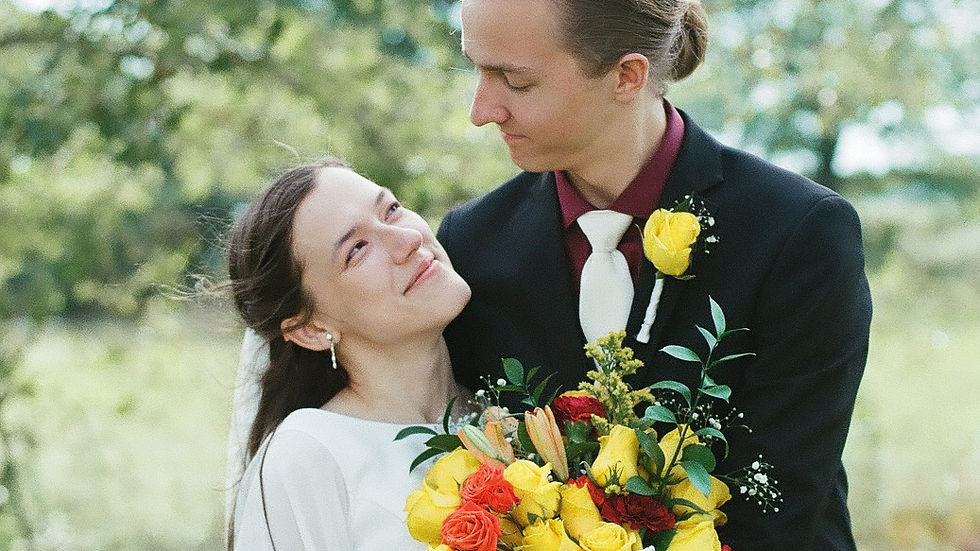 Bride and groom embrace, smiling at each other. Bride holds bouquet of yellow and red flowers. Lush green outdoor setting.
