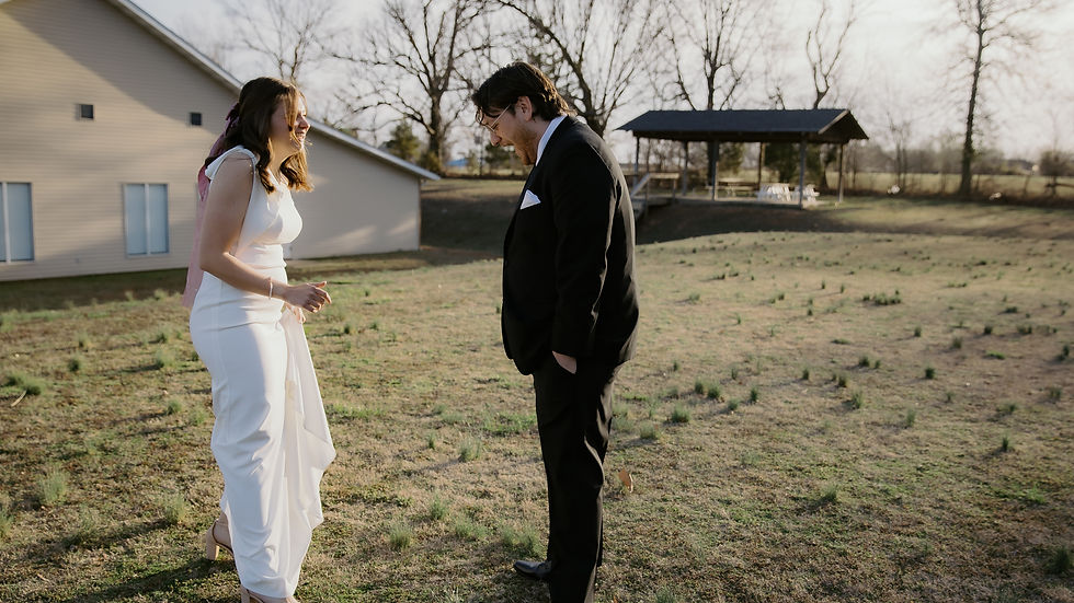 Bride in white dress and groom in black suit laughing in a yard with a house and trees in the background, creating a joyful scene.