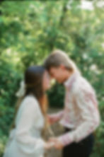 A couple in patterned shirts share a tender moment, foreheads touching, in a lush, sunlit forest, holding hands and smiling.