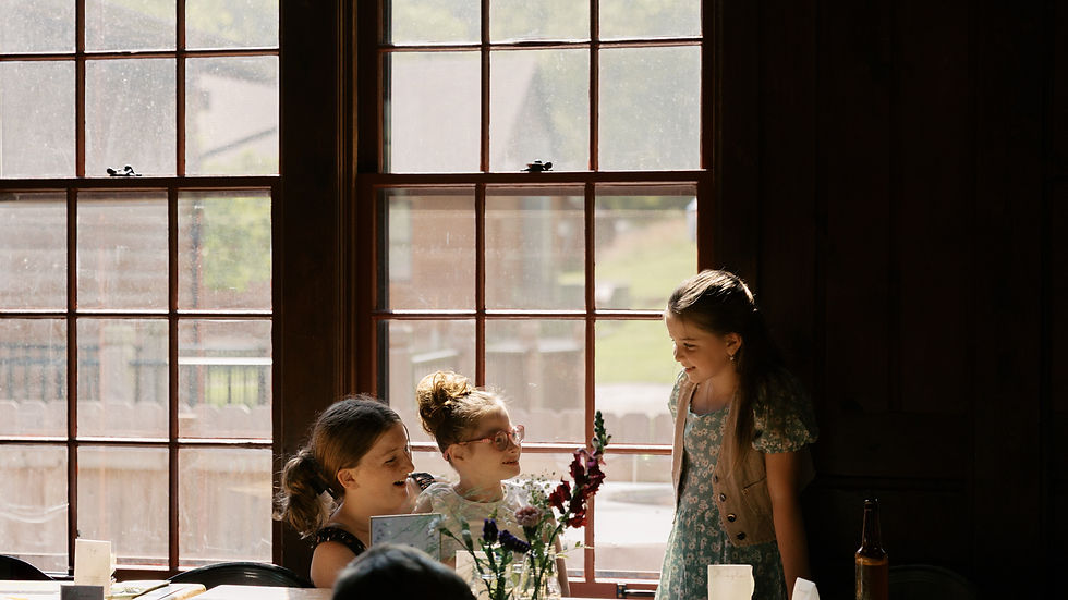 Three girls chat happily at a sunlit table by large windows. Flowers and name cards are on the table, creating a warm, cozy atmosphere.