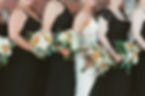 Bridesmaids in black dresses hold white and peach bouquets, standing beside a bride in a white gown. Brick background, cheerful mood.