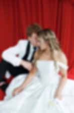 Couple in formal attire sits on a red background. The woman in a white dress, the man whispers affectionately. Elegant and romantic mood.