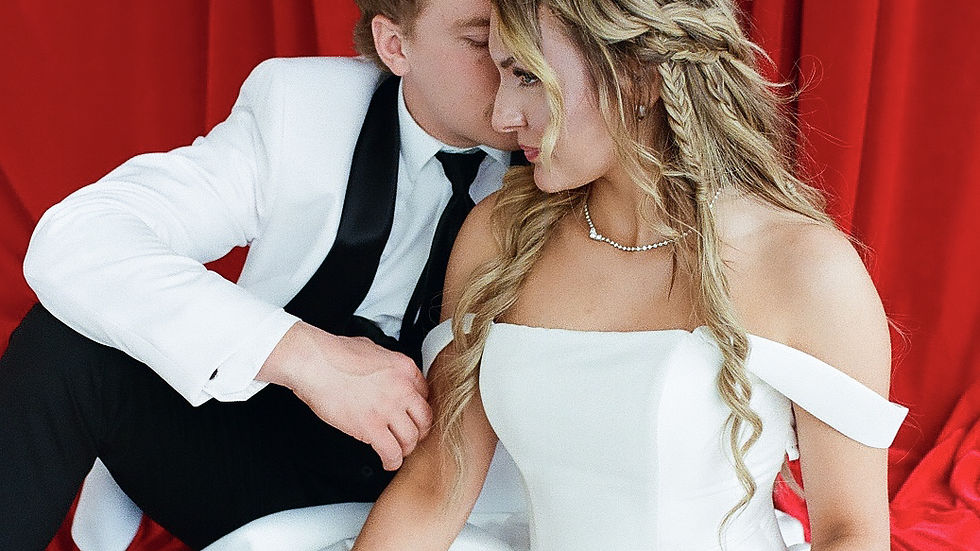 Couple in formal attire sits on a red background. The woman in a white dress, the man whispers affectionately. Elegant and romantic mood.