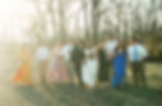 Bridal party walking outdoors, smiling. Bride in white, others in colorful dresses and suits, holding flowers. Sunlit, wooded background.