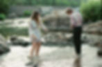Two people wading in a rocky creek, surrounded by greenery. The woman wears a light dress, the man a patterned shirt. Calm and serene setting.