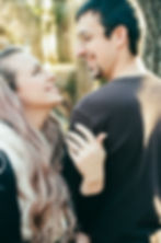 A couple smiling at each other outdoors, the woman touches the man's shoulder, displaying an engagement ring. Soft lighting, trees in background.