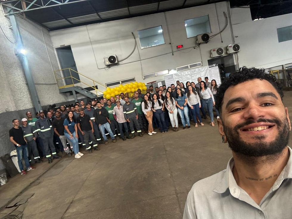 Homem faz selfie sorrindo com grande grupo de colegas em galpão, com balões amarelos ao fundo.