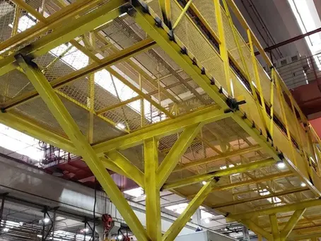 Passarela metálica amarela suspensa em ambiente industrial. Estrutura de malha visível no teto. Luzes e tubos ao fundo.