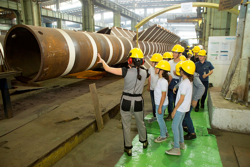 Crianças com capacetes amarelos visitam fábrica, guiadas por uma mulher de uniforme. Estrutura metálica ao fundo. Ambiente industrial.
