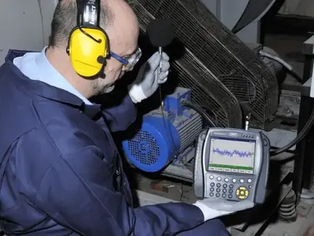 Técnico com fones amarelos mede ruído em máquina industrial, segurando aparelho com gráfico, em ambiente de manutenção.