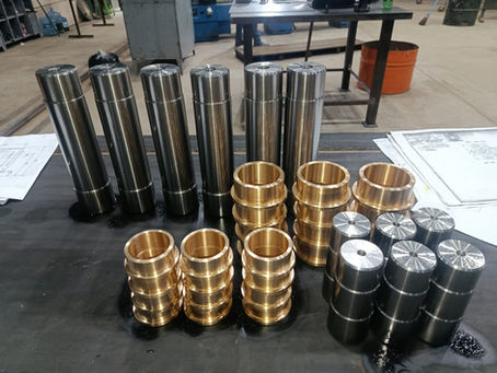 Peças metálicas usinadas em aço e latão sobre mesa de trabalho, com desenhos técnicos ao redor, em oficina industrial.