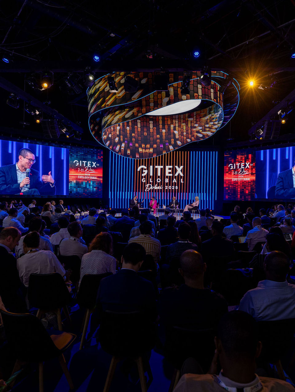 Wide shot of a massive technology exhibition auditorium featuring expansive, curved digital screens and a vast seated audience, photographed by a London exhibition photographer.