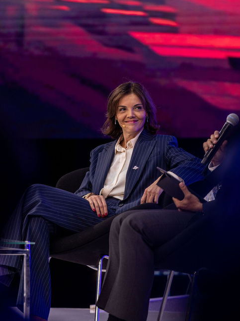 A female corporate executive in a pinstripe suit smiling during a stage panel discussion and fireside chat at a major business summit, photographed by a London corporate PR photographer.