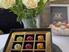 Brightly colourful jewel like chocolates created by Andrew King displayed within a luxury box with a gold AK logo, beautifully displayed on a white cloth with a vase of cream roses