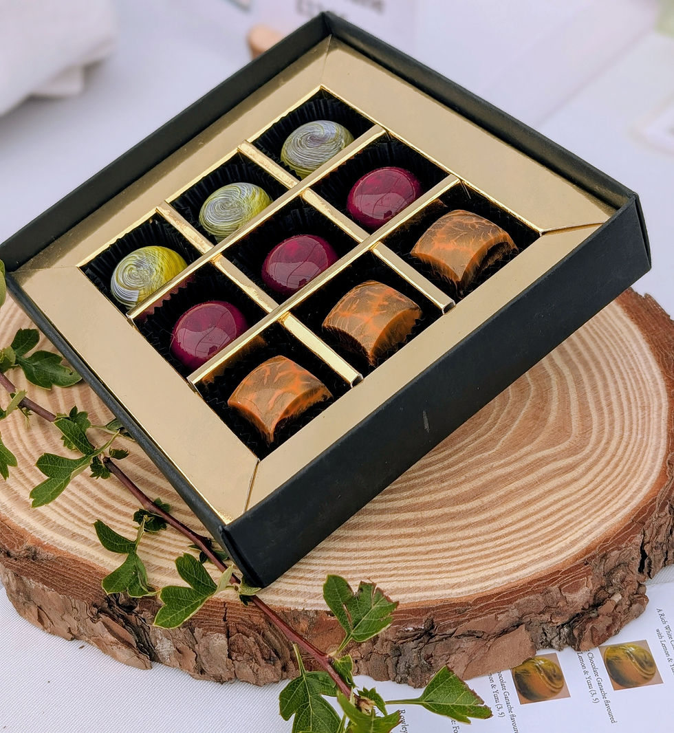 Box of colourful chocolates by Andrew King Chocolates with swirling patterns sits on a wooden slab with green leaves. Background features a white tablecloth.